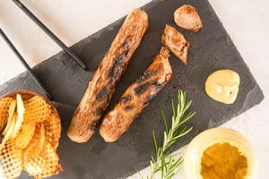 Grilled bratwurst on charcoal cutting board. Rosemary, chips and condiments on the side.