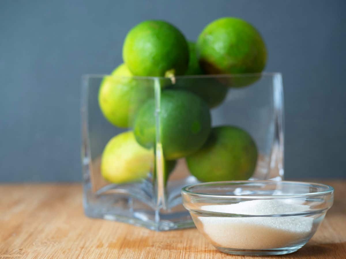 How to Candy Limes - Step 1 Limes in a glass container and sugar in a small bowl, wooden background.