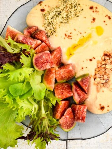Top view of hummus dip in a bowl with fresh lettuce, figs, nuts, hemp seeds and olive oil.