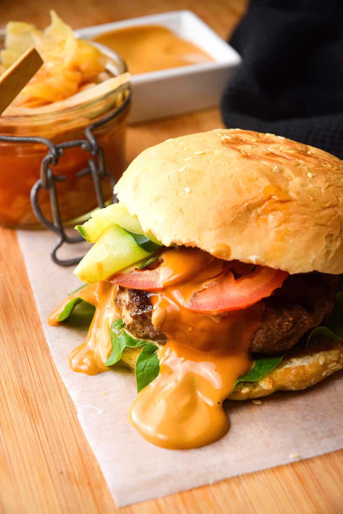 Kimchi burger with kimchi sauce on wooden background with sauce dish and a jar of kimchi on the side.