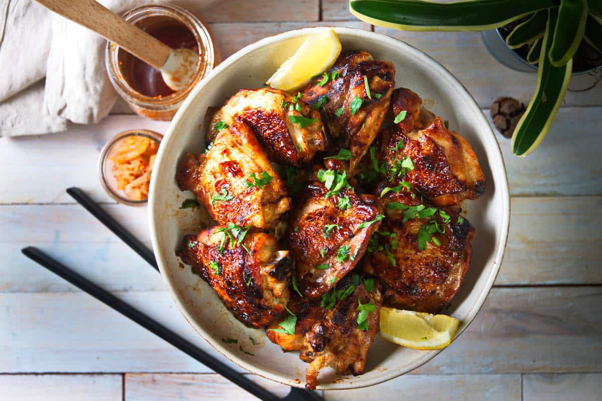 Kimchi chicken thighs and lemon wedges in dish. Chopsticks and kimchi on the side on white-washed wooden background.