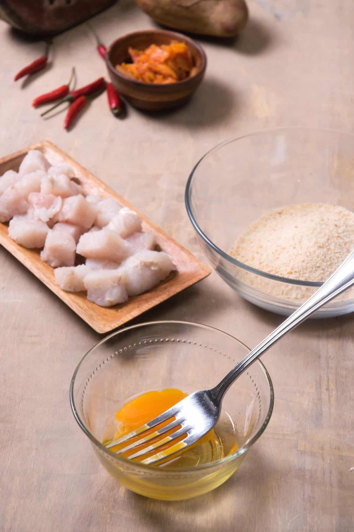 A small bowl of cracked eggs and a medium bowl with breadcrumbs., raw cod pieces on the side.
