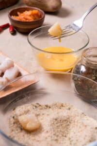 Raw cod pieces dipped in a small bowl of eggs beaten and a bowl of breadcrumbs.