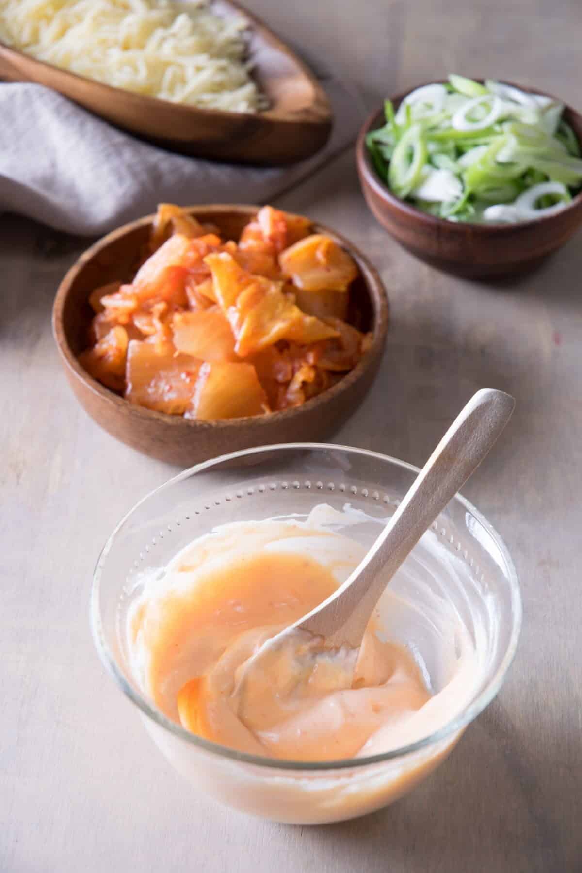Small bowls with creamy sriracha sauce, shredded cheese, kimchi and sliced scallions on light background.