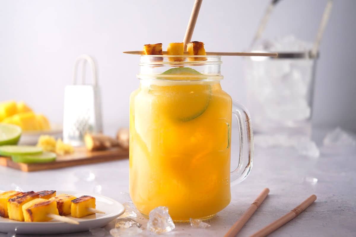 Mango ginger cocktail with grilled mango skewer and limes slices in a glass jar.