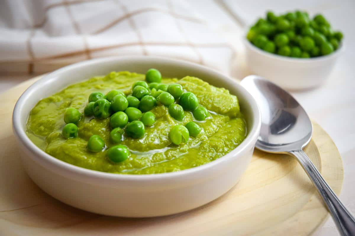 Pea purée in a white bowl on wooden plate, with a spoon on the side