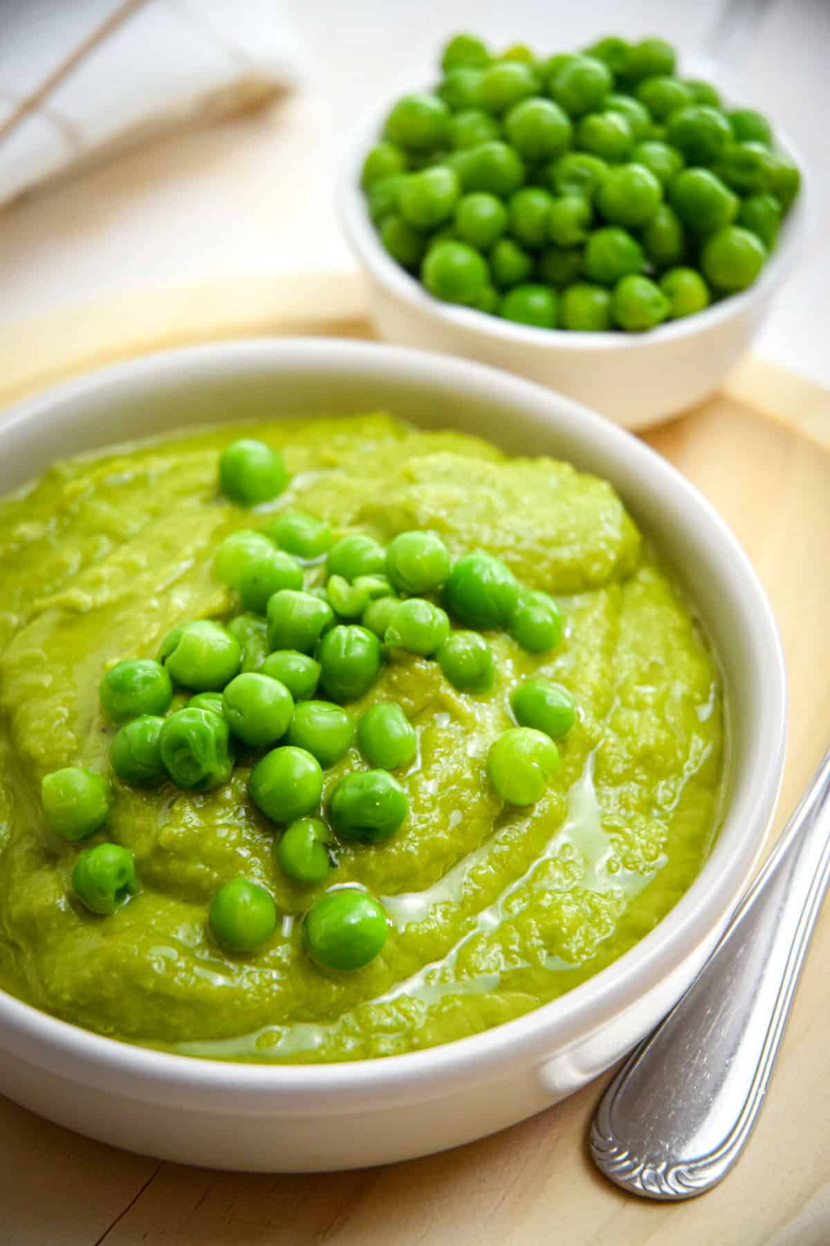 Pea purée in a white bowl on wooden plate, with a spoon on the side and green peas in the background.