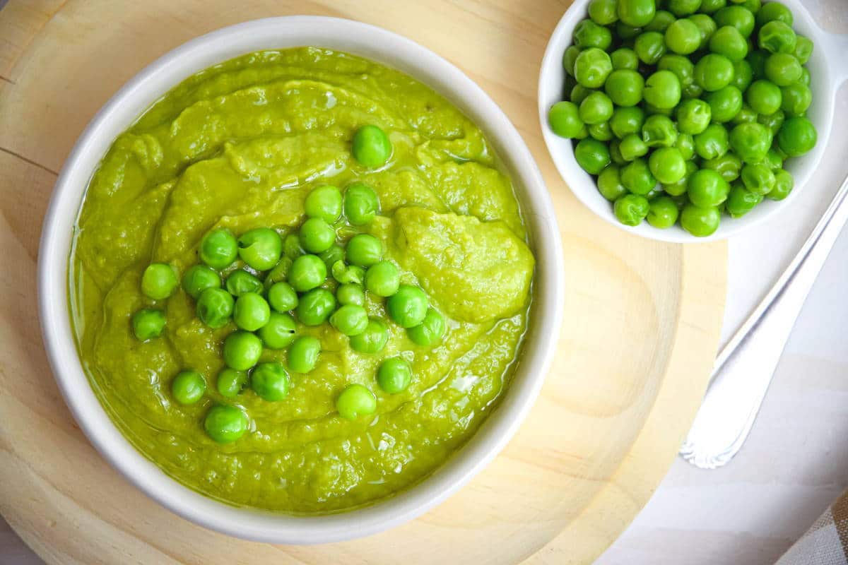 Pea purée in a white bowl on wooden plate, with green peas on the side.