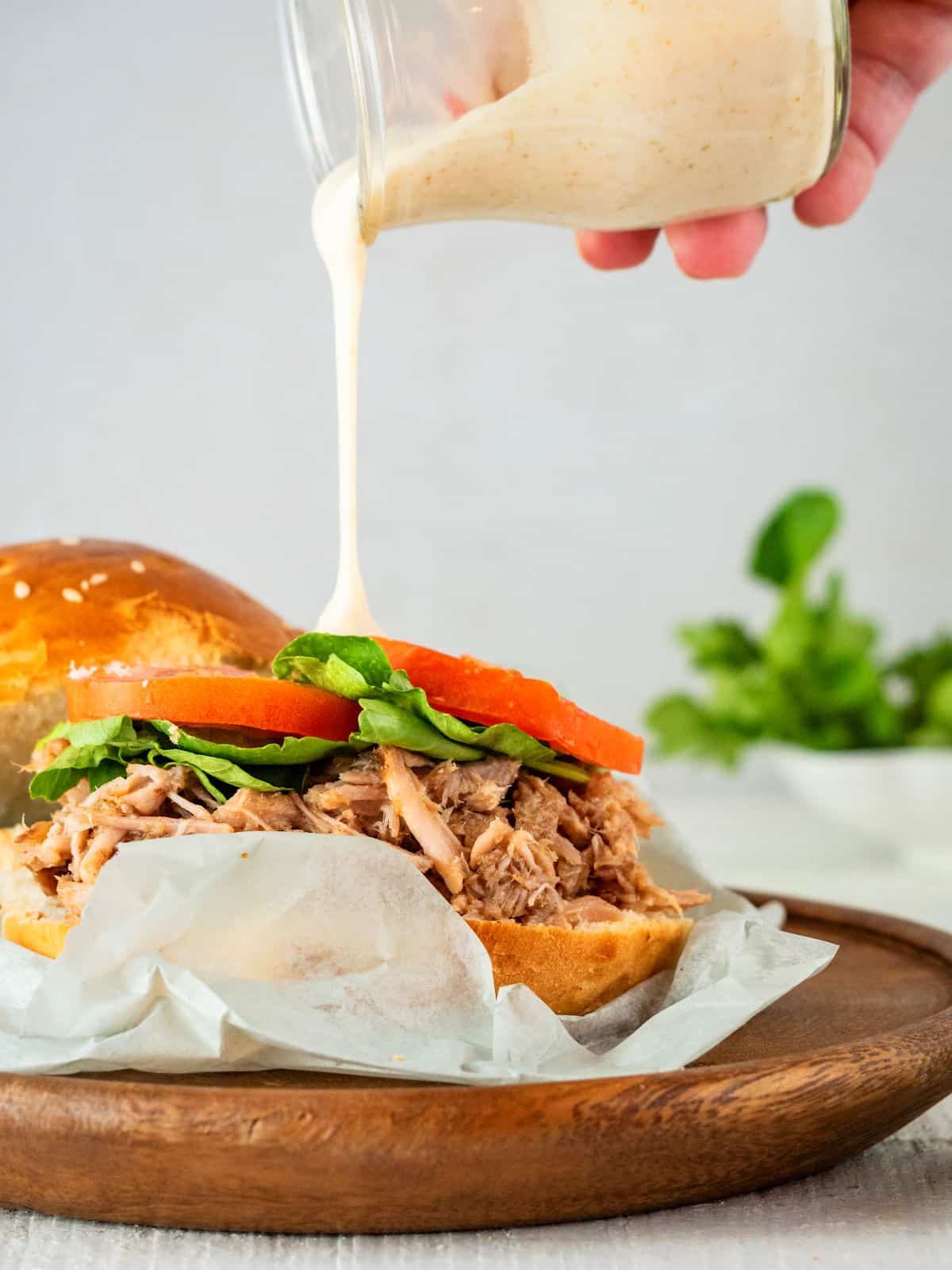 Pulled pork burger on wooden plate with special sauce drizzling over top.
