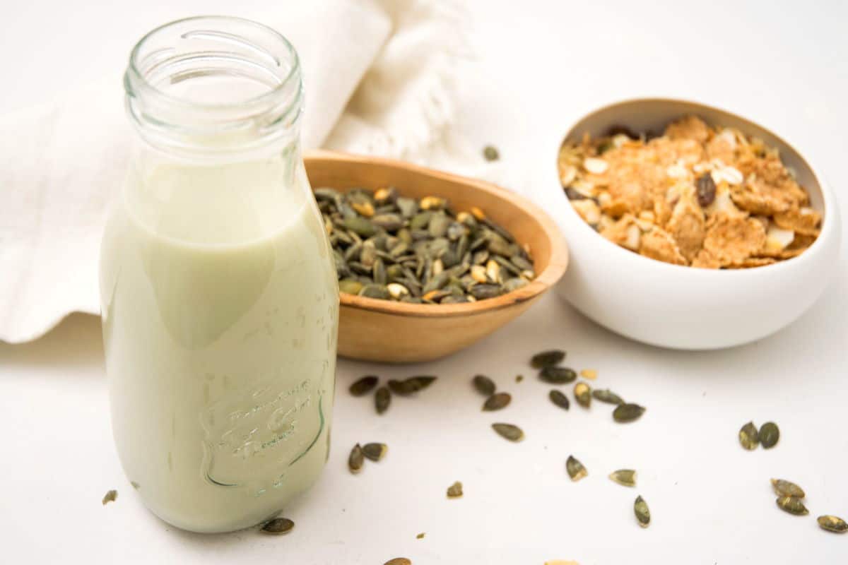 Pumpkin seed milk in a small jar on white background with pumpkin seeds in the background.