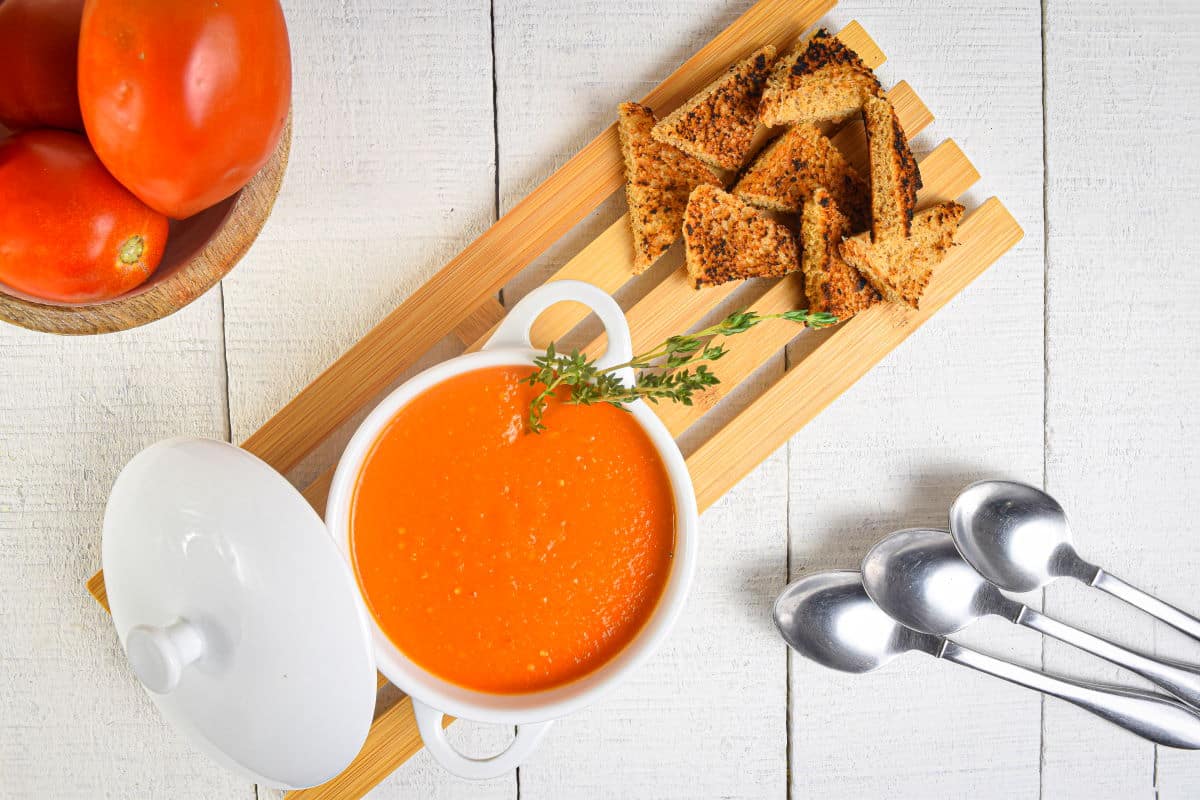 Roasted tomato soup in white bowl with toast points and spoons on the side.