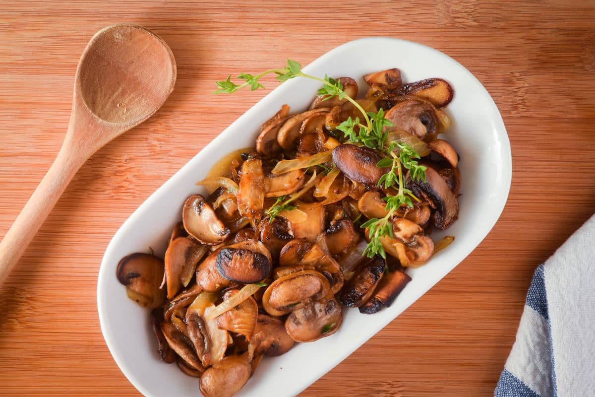 Sautéed mushrooms in white serving dish with fresh thyme, wooden spoon on the side.