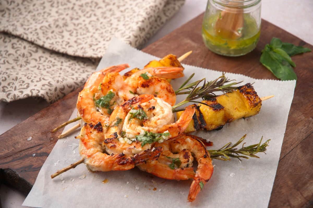 Shrimp on Rosemary Skewers with Rum-Soaked Pineapple Grilled shrimp on rosemary skewers and skewered pineapple wedges on parchment paper with sauce on the side.