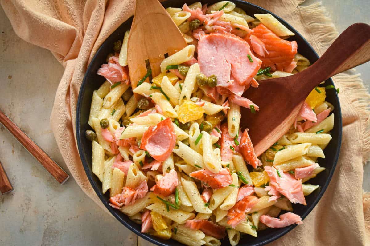Smoked salmon pasta salad in a dark bowl with a wooden spoon and off-white dish cloth.