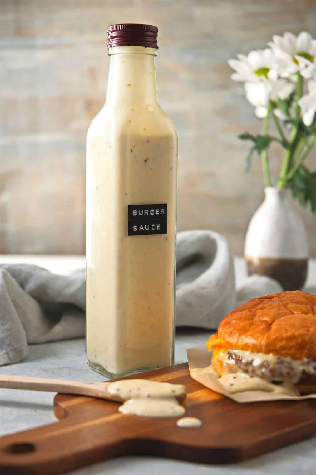 Burger sauce in a tall, labeled bottle with hamburger on the side and white flowers in the background.