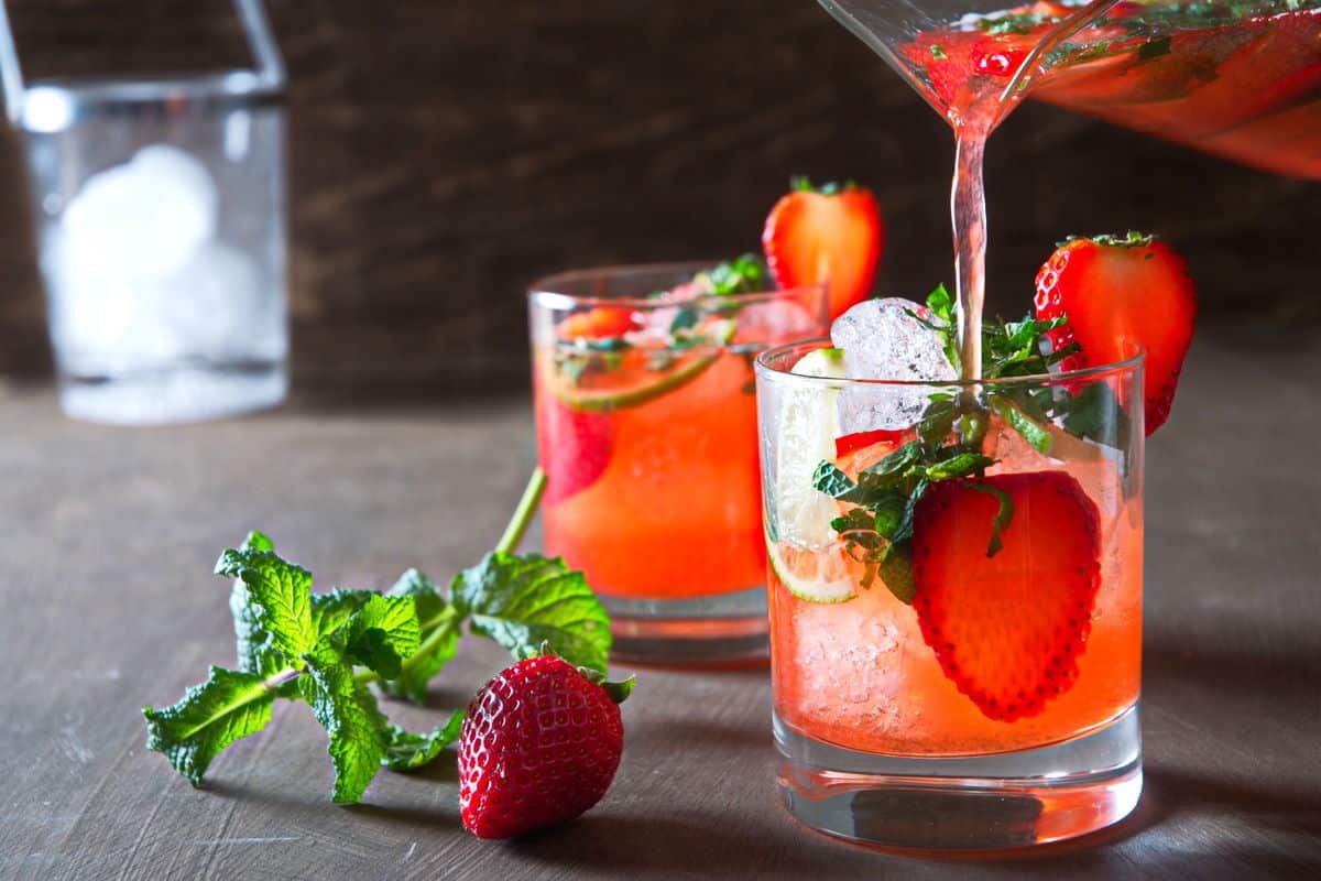 Strawberry mojito mocktail pouring out of a pitcher into glasses with fresh strawberries and mint on the side.