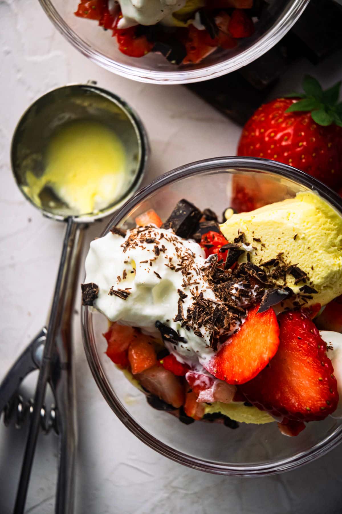 2 strawberry sundaes in dessert bowls, strawberries and ice cream scoop on the side.