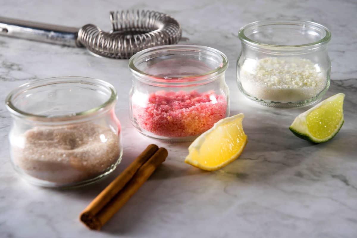 Sugar rims in 3 small jars, a lime wedge, lemon wedge and cinnamon stick on the side.