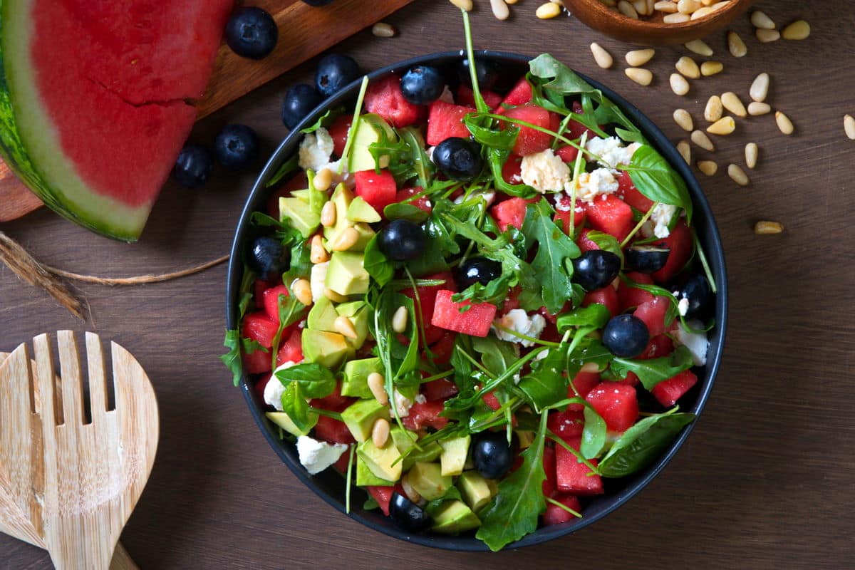 Watermelon salad in a black bowl on wooden background with salad spoons on the side.