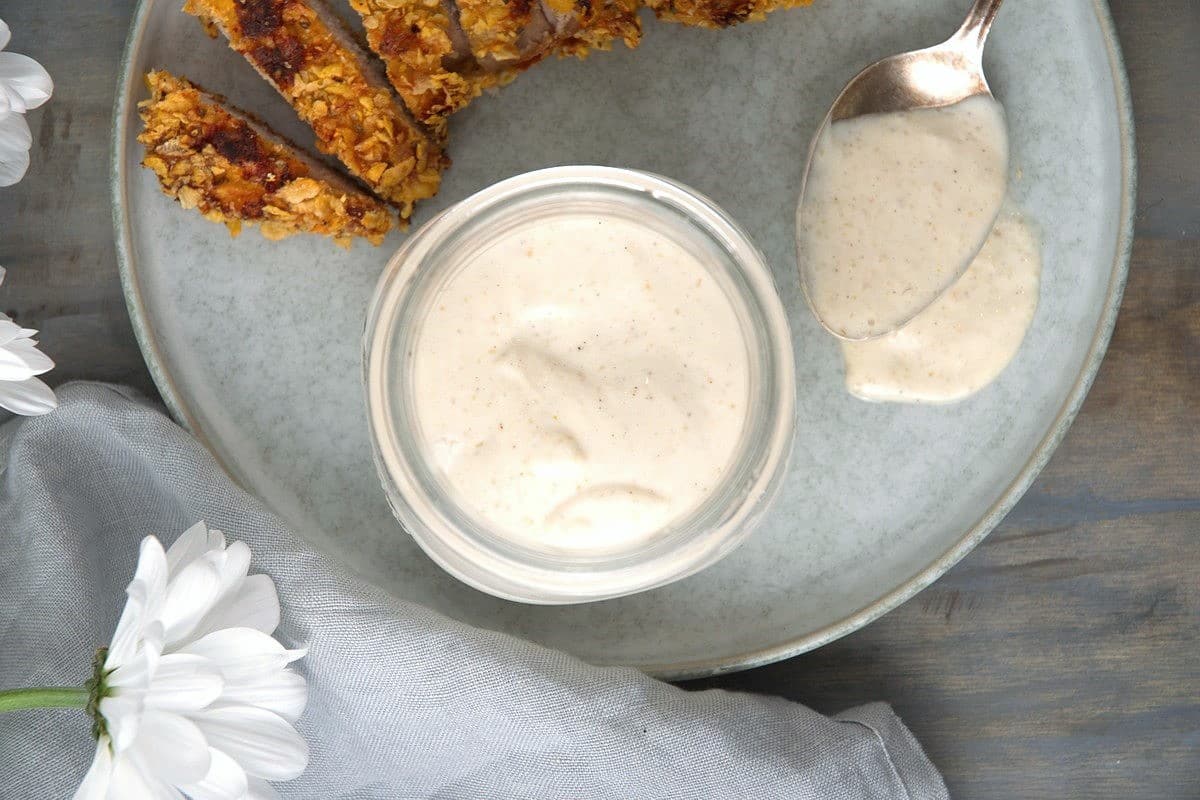 White BBQ sauce in a small jar on a plate with sliced, breaded chicken breast on the side.