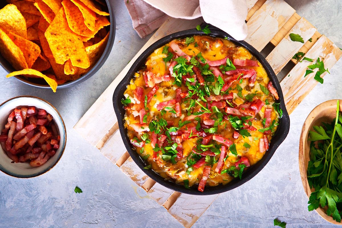Bison cheeseburger dip in a cast iron pan with tortilla chips and bacon bits on the side.