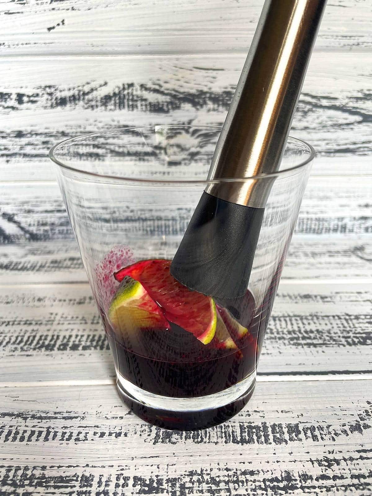 Lime wedges and blueberry syrup in glass with muddler.