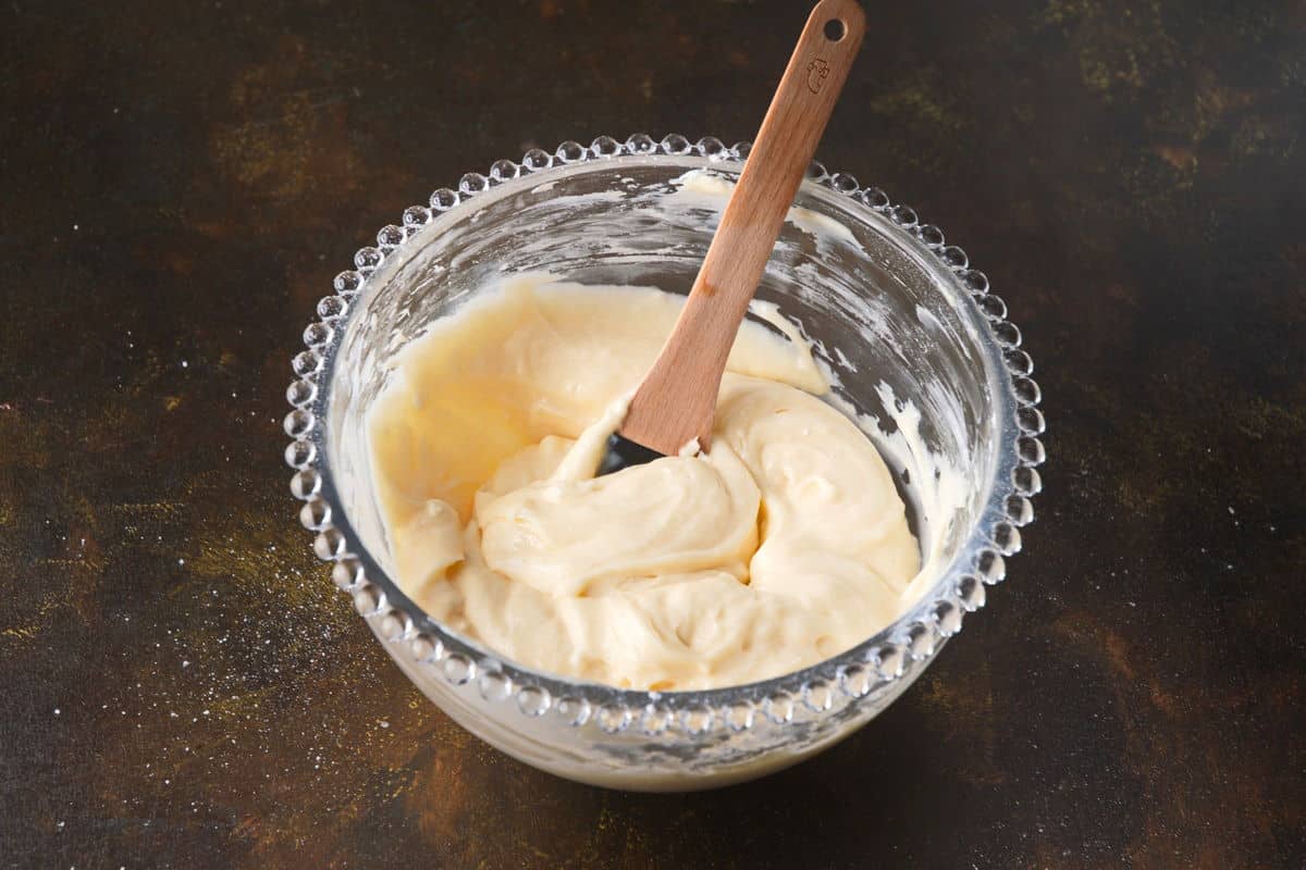 Cake pop frosting in bowl with spatula on brown background.