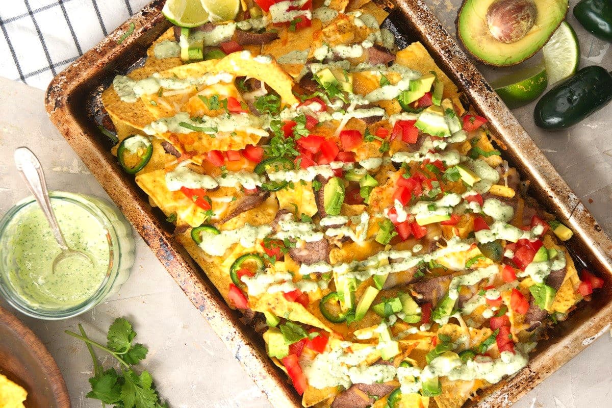 Carne asada nachos on sheet pan with an avocado, dip and jalapeños on the side.