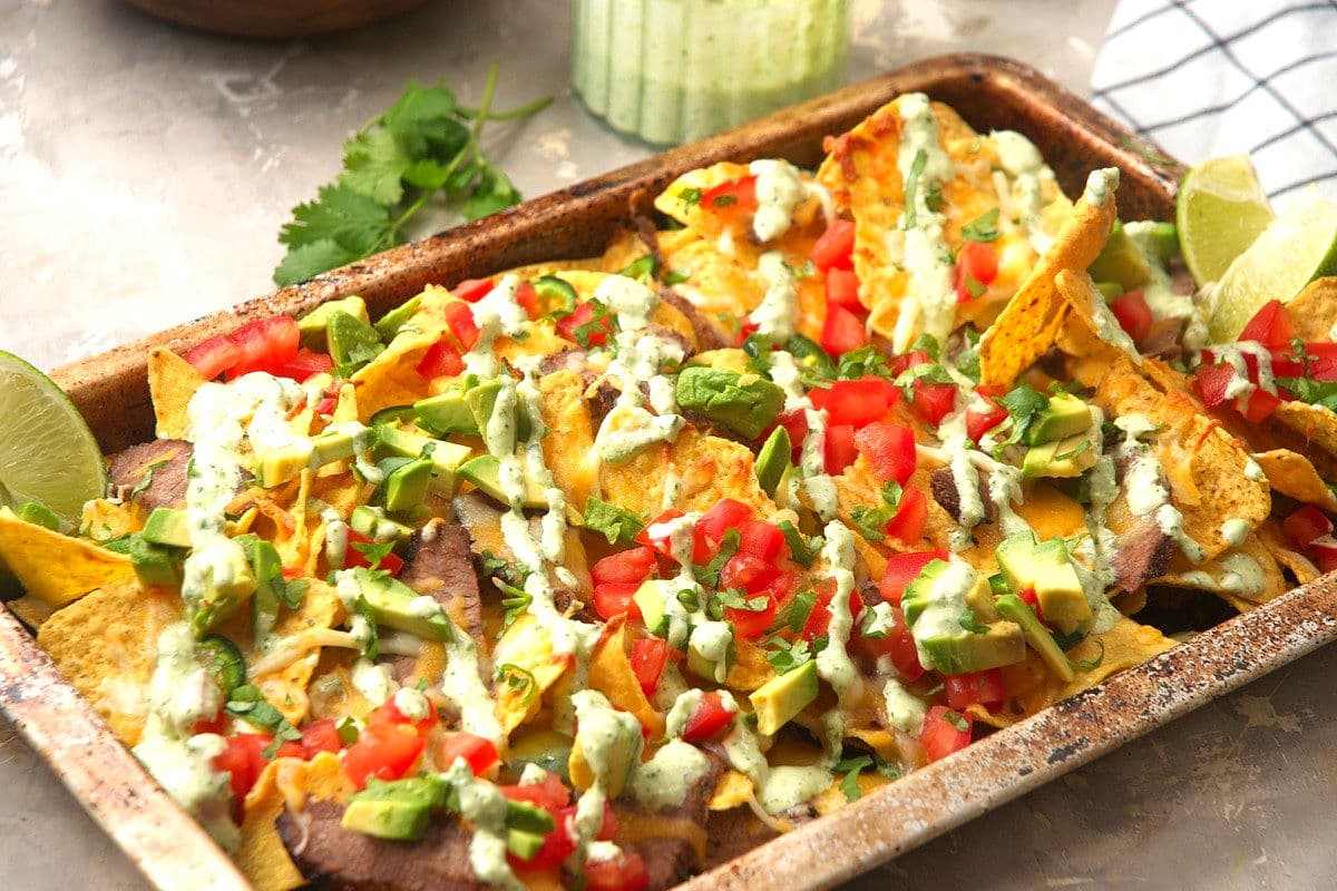 Carne asada nachos with creamy cilantro drizzle on baking sheet.