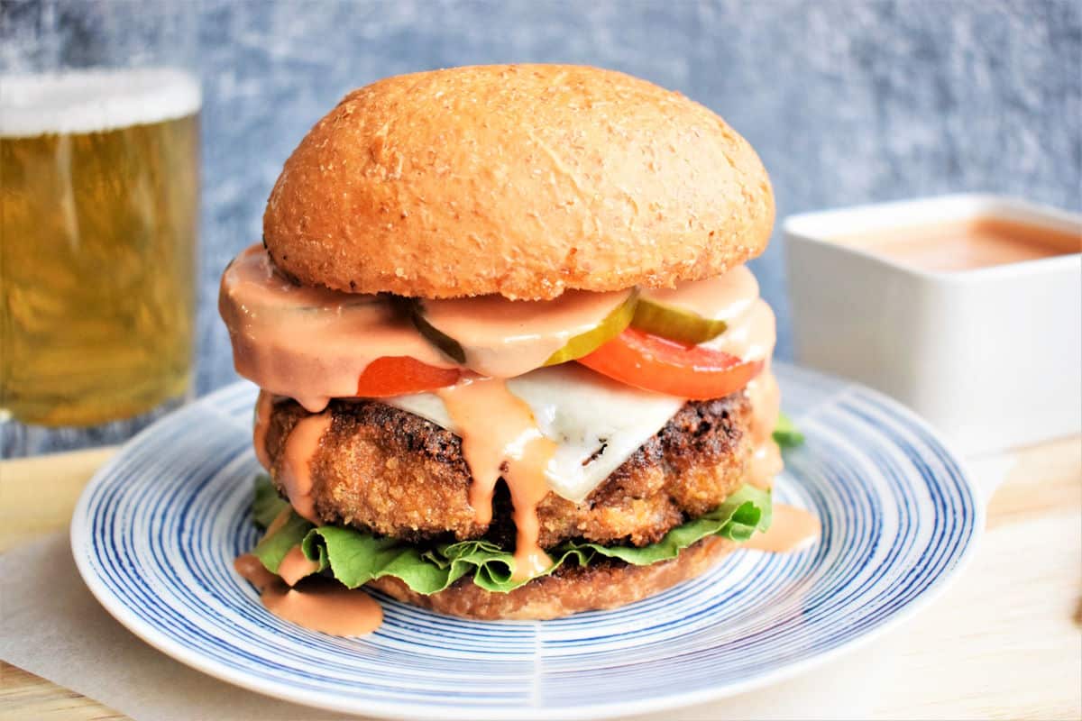 Chicken fried breaded hamburger on plate with beer and burger sauce in the background.