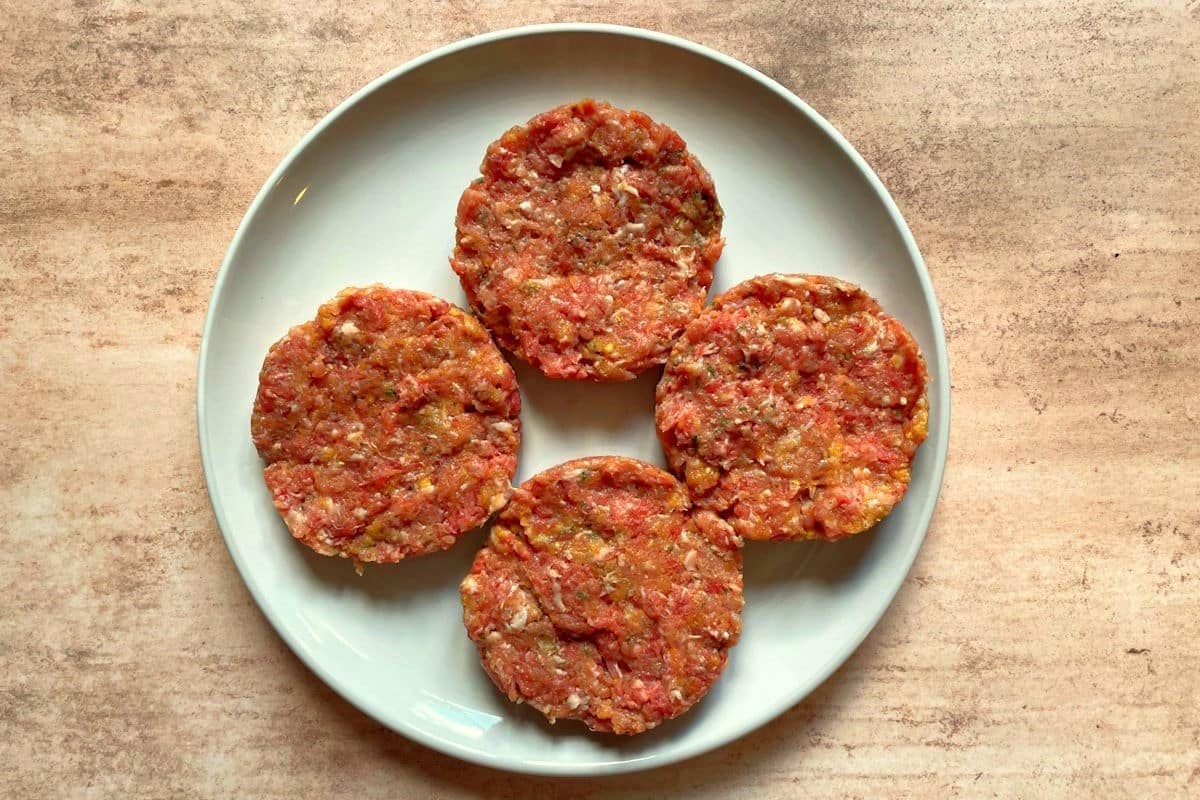 Raw chicken fried breaded burger patties on white plate.