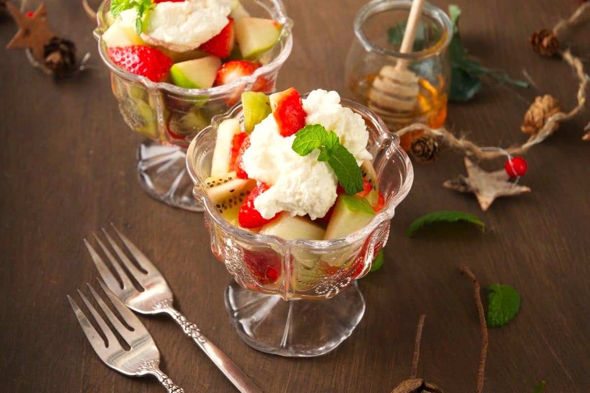 Fruit salad with whipped cream and mint in dessert glasses on brown background.