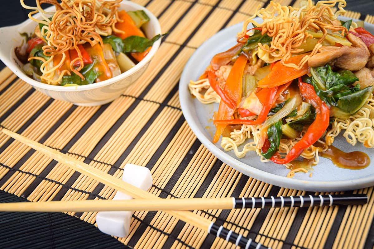 Crunchy chow mein noodle dish on white plate with chopsticks and wooden placemat on the side.