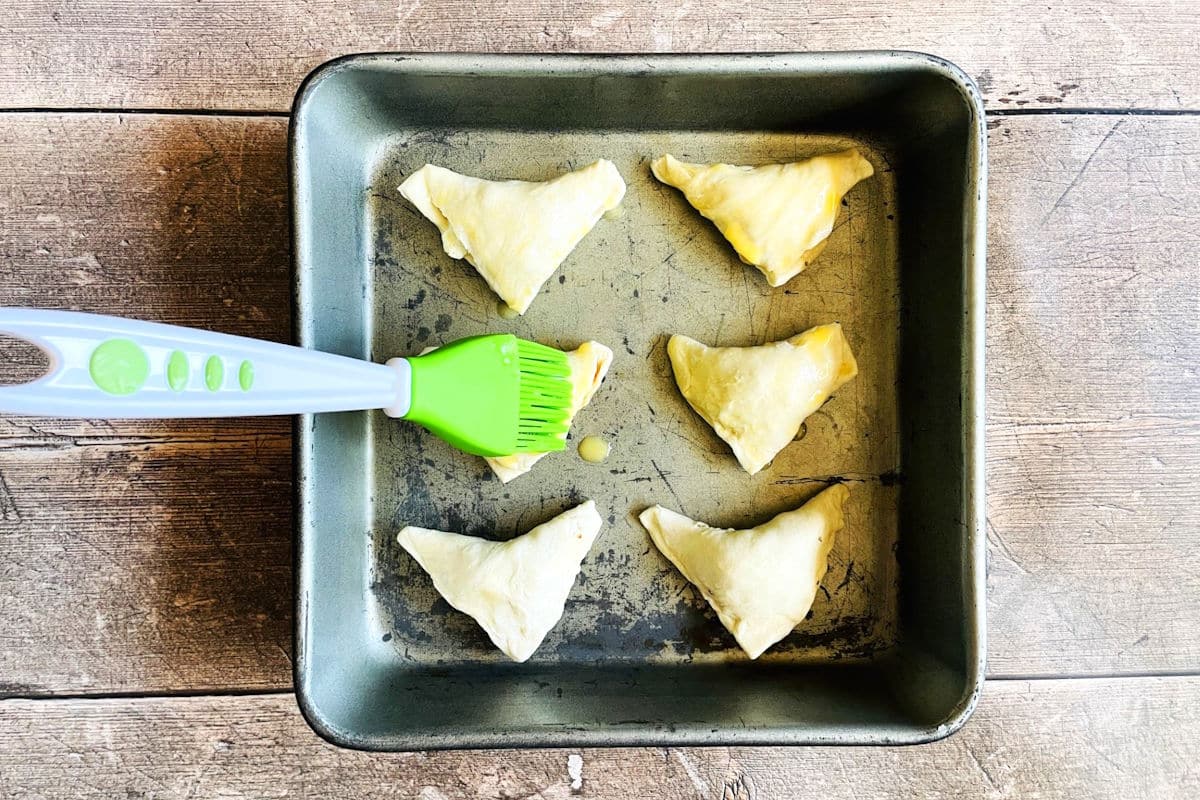 Puff pastry triangles in baking pan getting brushed with egg.