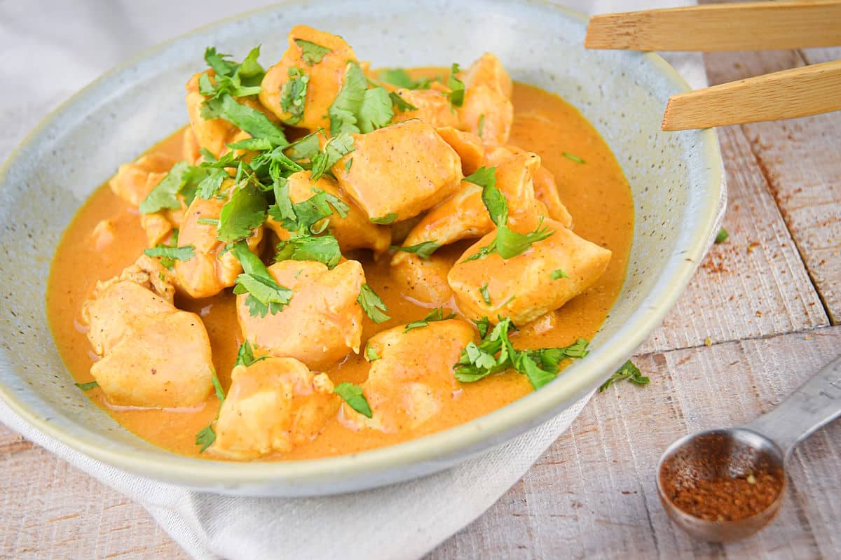 Butter chicken in a white specked bowl with wooden tongs on the side.