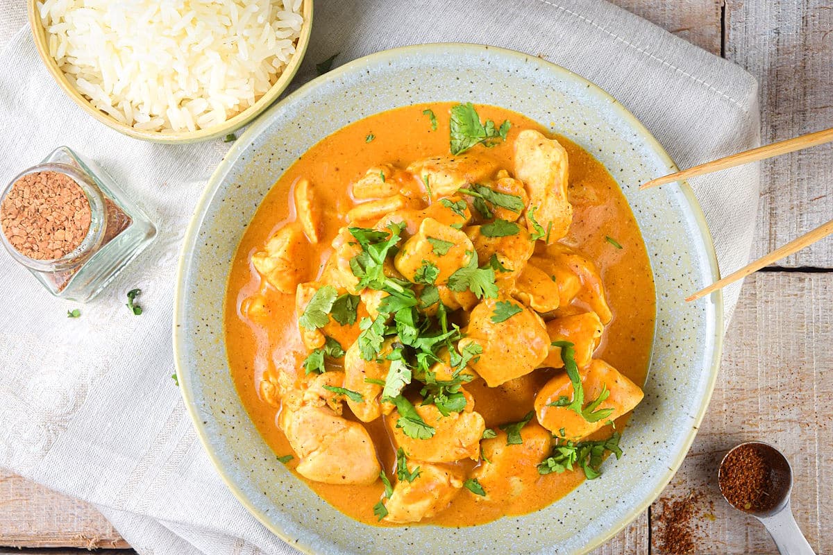 Butter chicken in a white specked bowl with wooden tongs, rice and spices on the side.