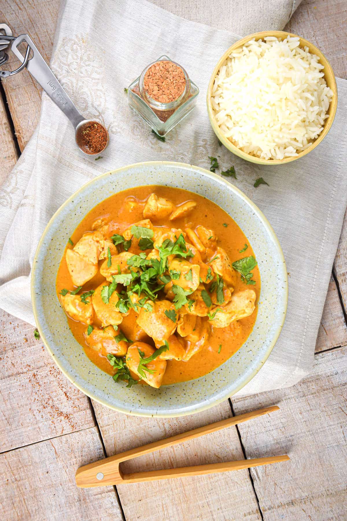 Butter chicken in a bowl with wooden tongs, rice and spices on the side.