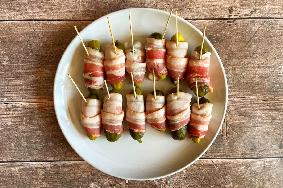 Raw bacon wrapped pickles with toothpicks on plate.