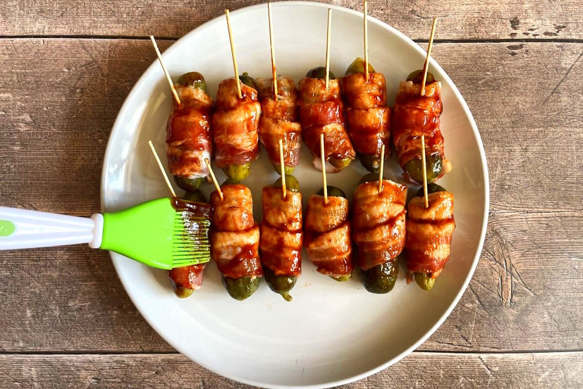 Raw bacon wrapped pickles with BBQ sauce on plate.