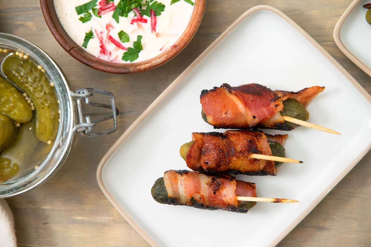 Bacon wrapped pickles on white serving plate with pickles on the side.