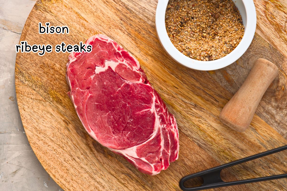 Raw bison ribeye steaks labeled and steak spice in a bowl on cutting board.