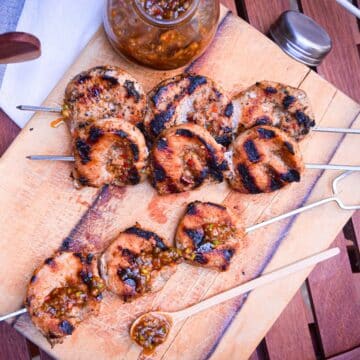 Grilled jerk pork tenderloin skewers on serving board with jerk sauce on the side.