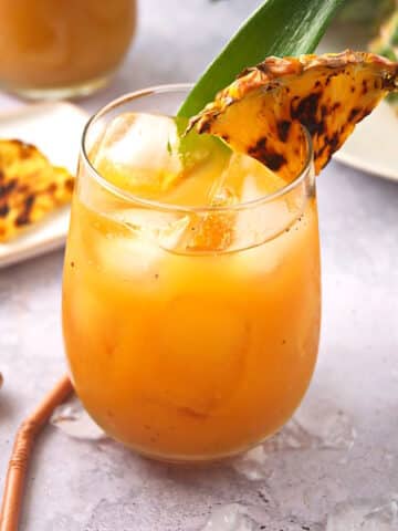 Grilled pineapple juice in stemless wine glass with grilled pineapple wedge garnish.