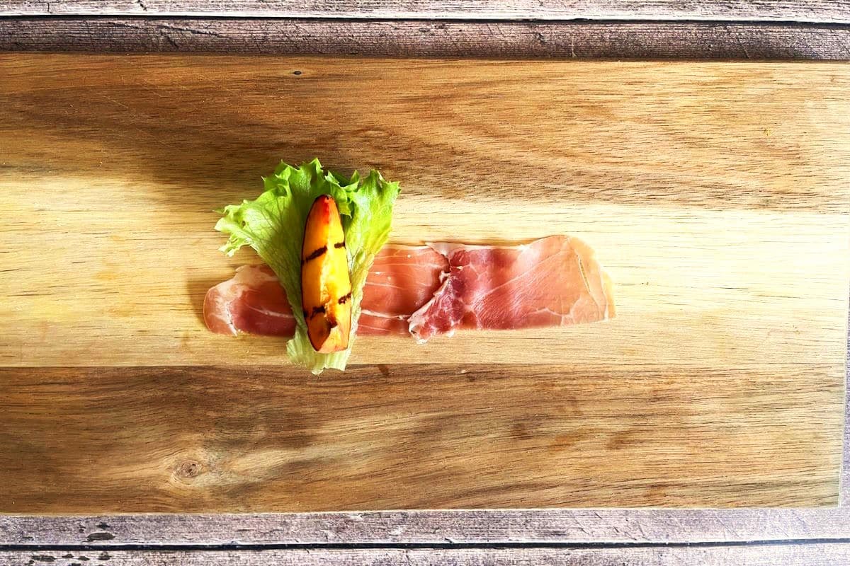 Grilled peach wedge, lettuce and prosciutto slice on cutting board.