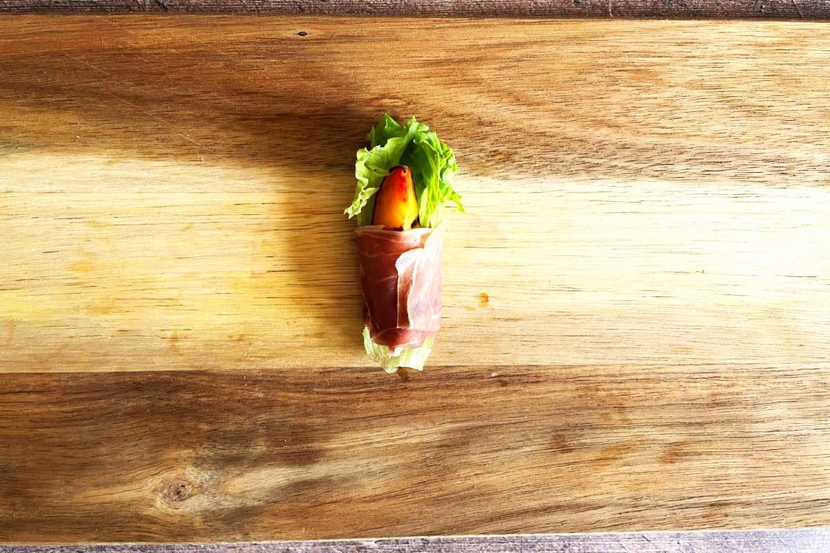 Grilled peach wedge with lettuce leaf wrapped up in prosciutto on cutting board.