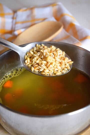 Uncooked pearl barley in spoon over a pot of soup.