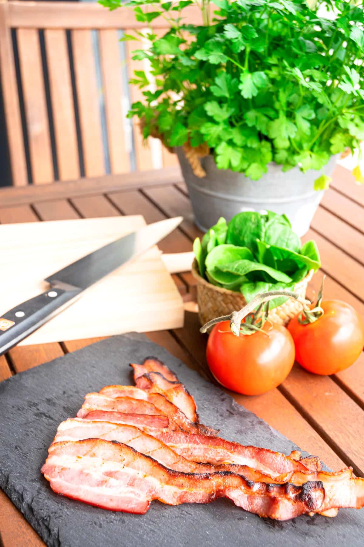 Grilled bacon on outdoor table with fresh herbs and tomatoes on the side.