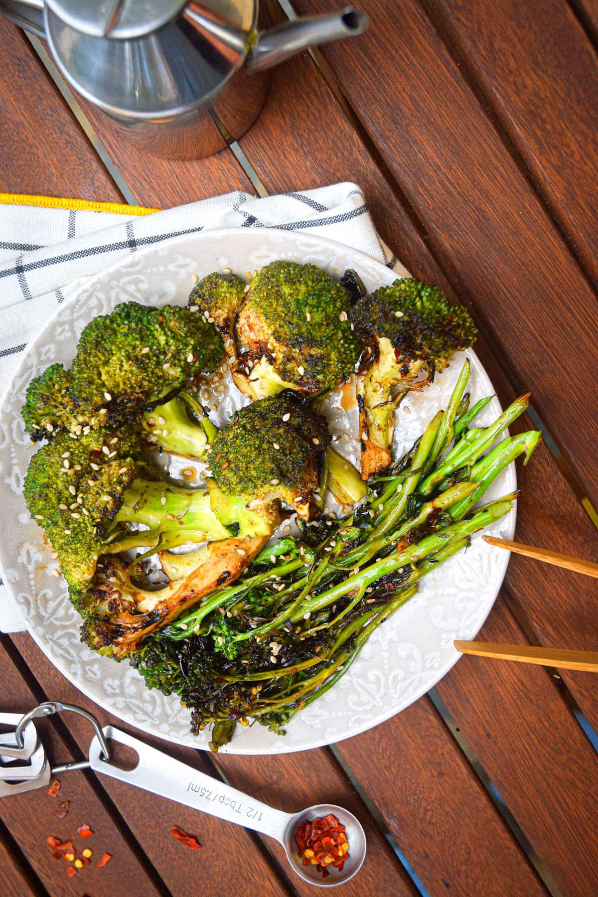 Grilled broccoli on plate on outdoor wooden table with red pepper flakes on the side.