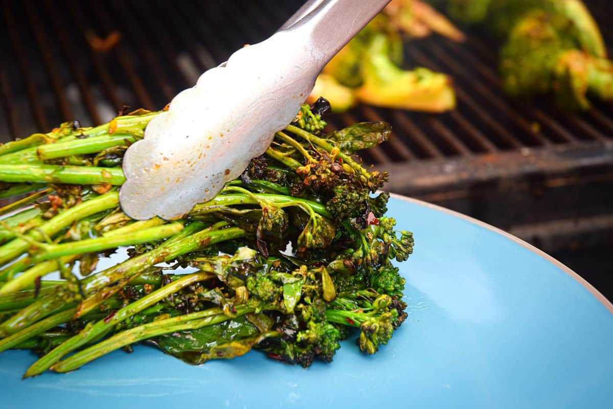 Grilled broccolini in tongs on blue plate.