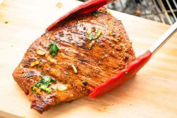 Grilled carne asada on cutting board in tongs.